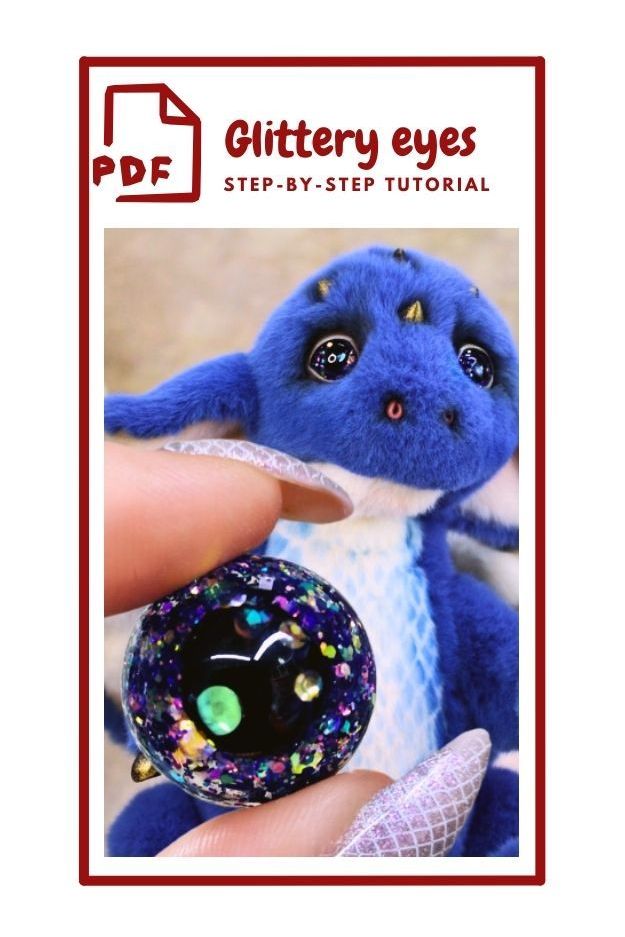 The image shows a cover for a PDF titled "Glittery eyes STEP-BY-STEP TUTORIAL." It features a close-up of a small, plush blue dragon with large, shiny eyes. In the foreground, a hand with long, glittery nails is holding a round, glittery object that resembles an eye, with colorful sparkles inside. The dragon's eyes and the object in the hand both have a glittery appearance. ___  PDF  Glittery eyes STEP- BY- STEP TUTORIAL   text, animal, toy, cartoon, handmade. a hand holding a stuffed animal.